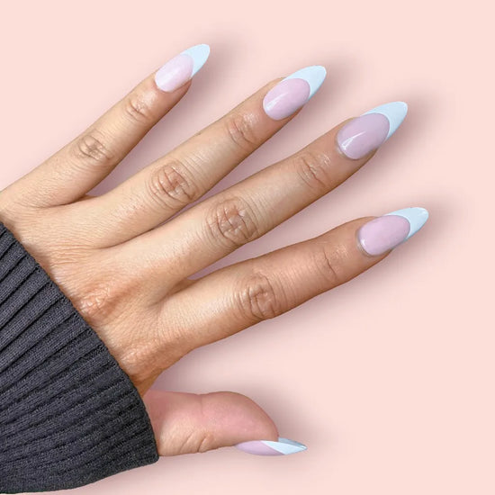 Diagonal spread hand showing de blinkie Lady Primrose soft sheer pink base with bold white French tip press-on nails in medium almond shape against soft pink background with dark charcoal ribbed sleeve