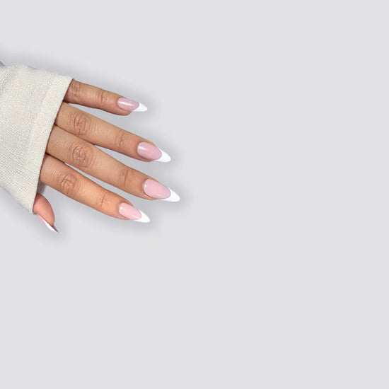  Spread hand showing de blinkie Lady Primrose soft sheer pink base with bold white French tip press-on nails in medium almond shape against light grey background with natural linen sleeve