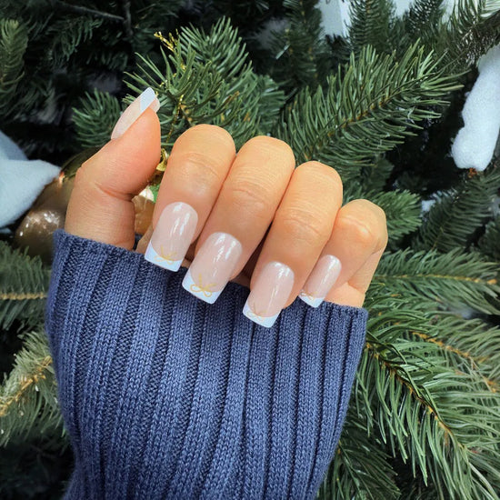 Claw pose showing de blinkie Luxe Ribbon French sheer nude base with white French tip and gold bow nail art in short square shape against Christmas tree background with blue ribbed sleeve