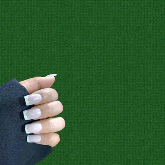 Claw pose showing de blinkie Luxe Ribbon French sheer nude base with white French tip and gold bow nail art in short square shape against deep green background with dark sleeve