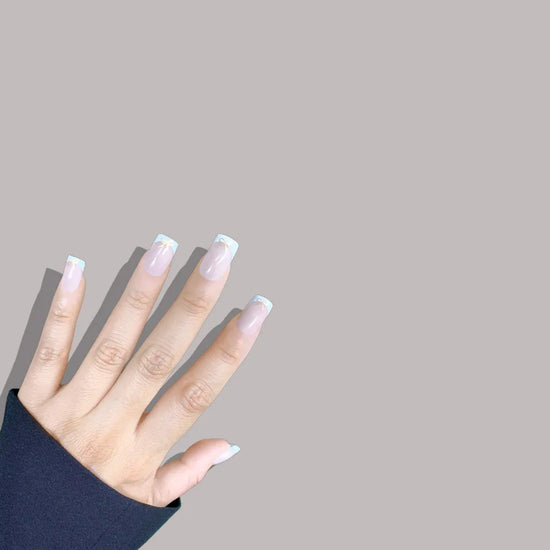 Spread hand showing de blinkie Luxe Ribbon French sheer nude base with white French tip and gold bow nail art in short square shape against warm grey background with dark sleeve