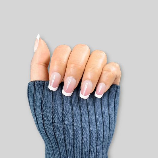 Downward claw pose showing de blinkie Madame Grace sheer pink white French tip press-on nails in short square against a soft grey background with a blue ribbed knit sleeve
