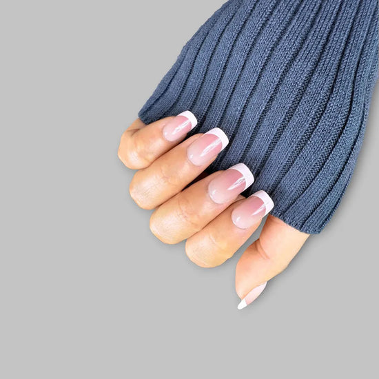 Upward claw pose showing de blinkie Madame Grace sheer pink white French tip press-on nails in short square against a soft grey background with a blue ribbed knit sleeve