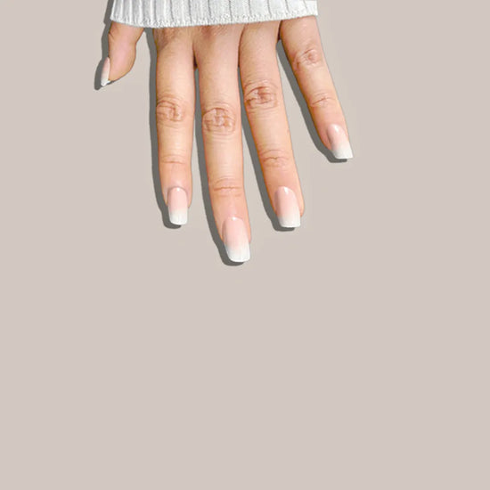 Downward spread hand showing de blinkie Mademoiselle Glow soft peach nude to white ombre press-on nails in short square against a warm taupe background with a white ribbed knit sleeve