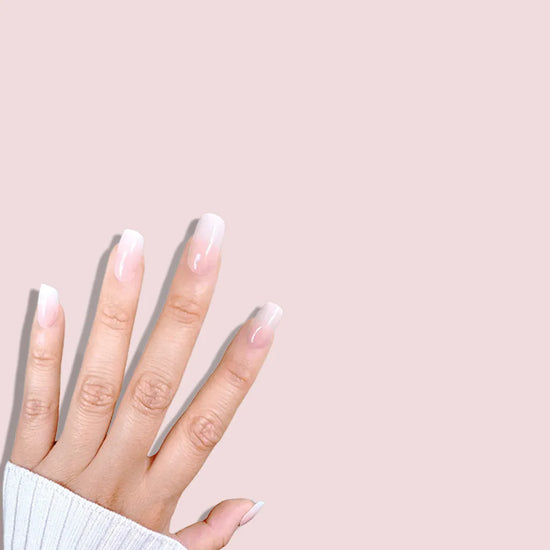 Spread hand showing de blinkie Mademoiselle Glow soft peach nude to white ombre press-on nails in short square against a soft pink background with a white ribbed knit sleeve