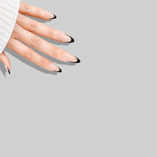 Diagonal downward spread hand showing de blinkie Midnight Frenchie soft nude base with black French tip press-on nails in short almond against a soft grey background with a white ribbed knit sleeve