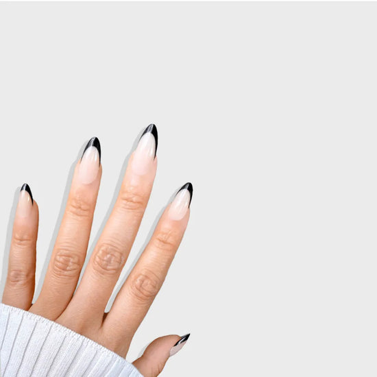 Spread hand showing de blinkie Midnight Frenchie soft nude base with black French tip press-on nails in short almond against a soft light grey background with a white ribbed knit sleeve