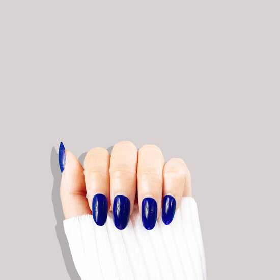 Upward claw pose showing de blinkie Midnight Reign cobalt blue gloss press-on nails in short oval against a grey background with a white ribbed knit sleeve