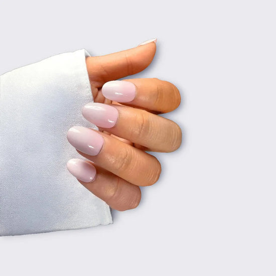 Claw pose showing de blinkie Milky Kiss soft milky pink press-on nails in short oval shape against light grey background with white fabric sleeve