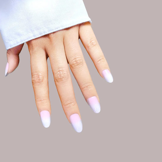 Down-facing hand pose showing de blinkie Milky Kiss soft milky pink press-on nails in short oval shape against warm grey background with white sleeve
