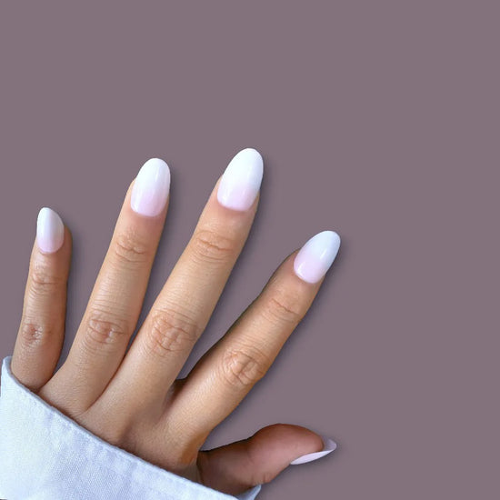Flat hand pose showing de blinkie Milky Kiss soft milky pink press-on nails in short oval shape against muted mauve background with white sleeve