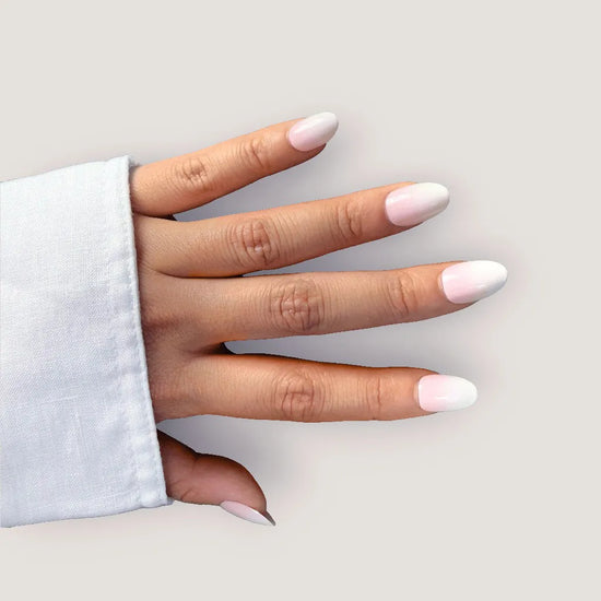 Spread hand showing de blinkie Milky Kiss soft milky pink press-on nails in short oval shape against light grey background with white linen sleeve