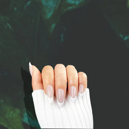 Claw pose showing de blinkie Miss Classé sheer blush base with white French tip press-on nails in short almond against a dark moody teal background with a white ribbed knit sleeve