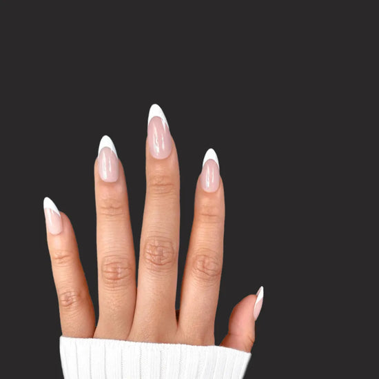 Diagonal upward spread hand showing de blinkie Miss Classé sheer blush base with white French tip press-on nails in short almond against a black background with a white ribbed knit sleeve
