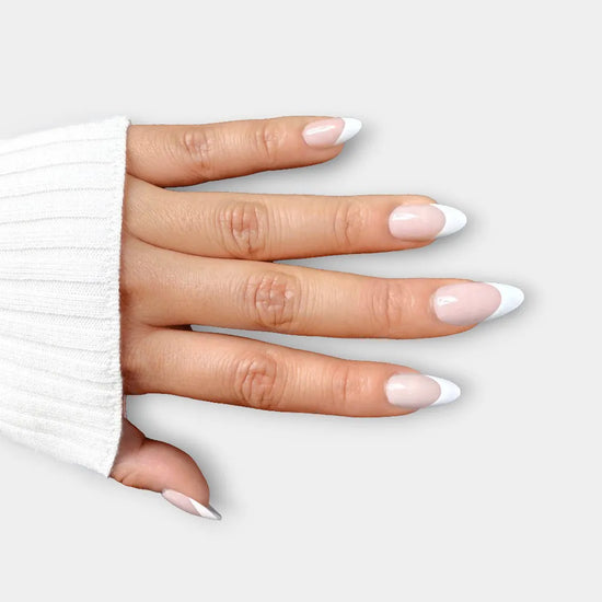 Spread hand showing de blinkie Miss Classé sheer blush base with white French tip press-on nails in short almond against a soft light grey background with a white ribbed knit sleeve