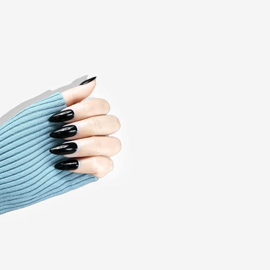 Claw pose showing de blinkie Miss Noir onyx gloss black press-on nails in short almond against a white background with a powder blue ribbed knit sleeve
