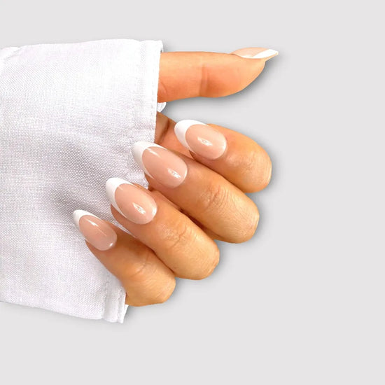 Tilted claw pose showing de blinkie Parisienne Blanc soft nude and white French tip press-on nails in short almond against a light grey background with a white linen cuff