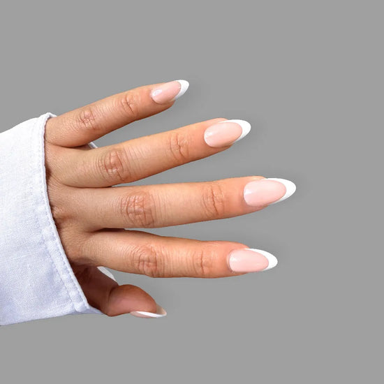 Spread hand showing de blinkie Parisienne Blanc soft nude and white French tip press-on nails in short almond against a grey background with a white shirt cuff