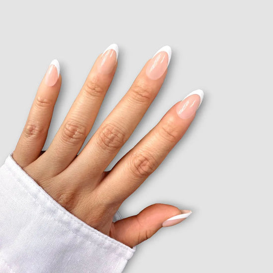 Upward-facing hand showing de blinkie Parisienne Blanc soft nude and white French tip press-on nails in short almond against a light grey background with a white shirt cuff