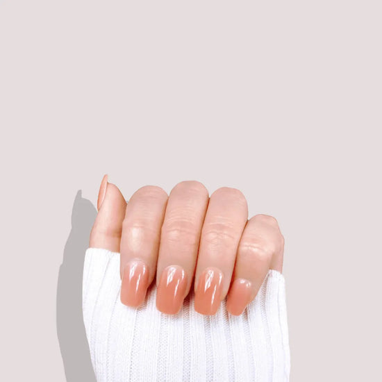 Claw pose showing de blinkie Peach Glow soft peach nude jelly press-on nails in short square against a soft blush pink background with a white ribbed knit sleeve