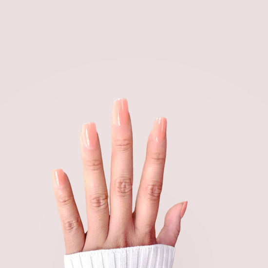 Fingers spread upward showing de blinkie Peach Glow soft peach nude jelly press-on nails in short square against a soft blush pink background with a white ribbed knit sleeve