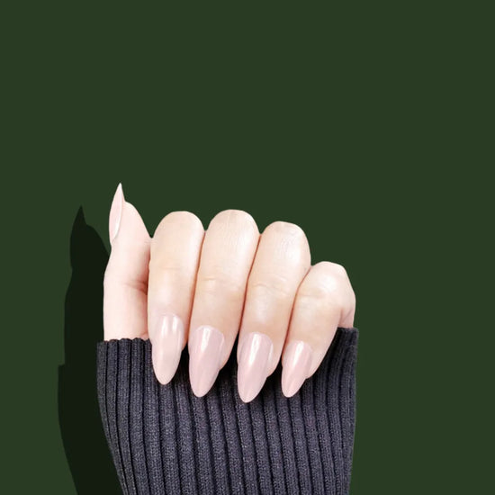 Claw pose showing de blinkie Peach Pearl soft peach nude pearl sheen press-on nails in short almond against a deep forest green background with a charcoal ribbed knit sleeve