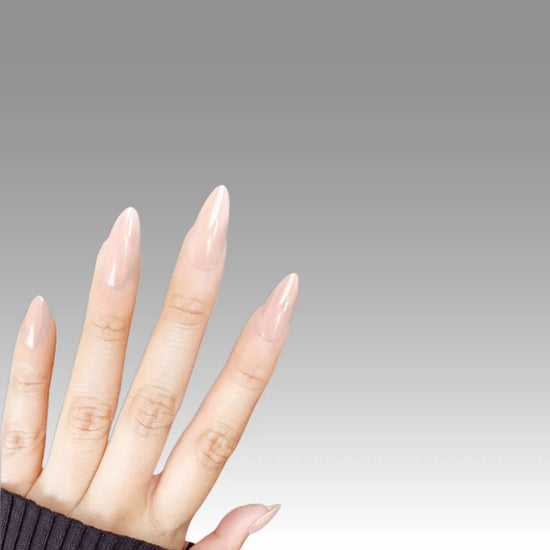 Diagonal left hand showing de blinkie Peach Pearl soft peach nude pearl sheen press-on nails in short almond against a grey gradient background with a charcoal ribbed knit sleeve