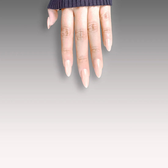 Fingers pointing downward showing de blinkie Peach Pearl soft peach nude pearl sheen press-on nails in short almond against a grey gradient background with a charcoal ribbed knit sleeve