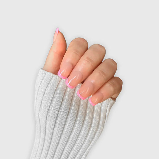 Claw pose showing de blinkie Pink Pop Frenchie sheer nude base with bubblegum pink French tip press-on nails in extra short square against a soft light grey background with a white ribbed knit sleeve