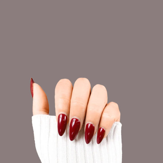 Claw pose showing de blinkie Plum Rouge deep plum burgundy gloss press-on nails in short almond against a grey background with a white ribbed knit sleeve