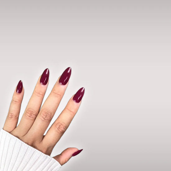 Diagonal hand pose showing de blinkie Plum Rouge deep plum burgundy gloss press-on nails in short almond against an off-white background with a white ribbed knit sleeve