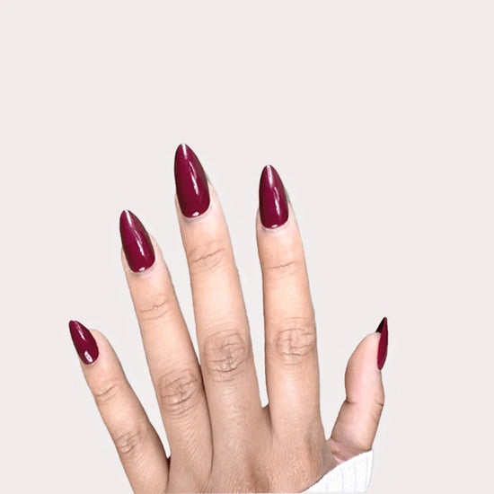 Fingers spread pose showing de blinkie Plum Rouge deep plum burgundy gloss press-on nails in short almond against an off-white background with a white ribbed knit sleeve