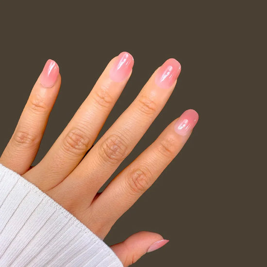Side angle hand pose showing de blinkie Rosé Jelly sheer warm rose ombre jelly press-on nails in extra short squoval shape against dark olive background with white ribbed sleeve