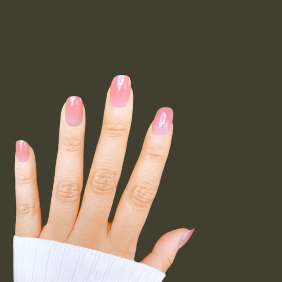 Spread hand showing de blinkie Rosé Jelly sheer warm rose ombre jelly press-on nails in extra short squoval shape against dark olive background with white ribbed sleeve