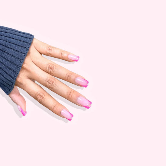 Diagonal hand pose showing de blinkie Rosé Mirage sheer blush pink with magenta French tips gold trace and 3D liner press-on nails in medium square shape against pink background with denim knit sleeve