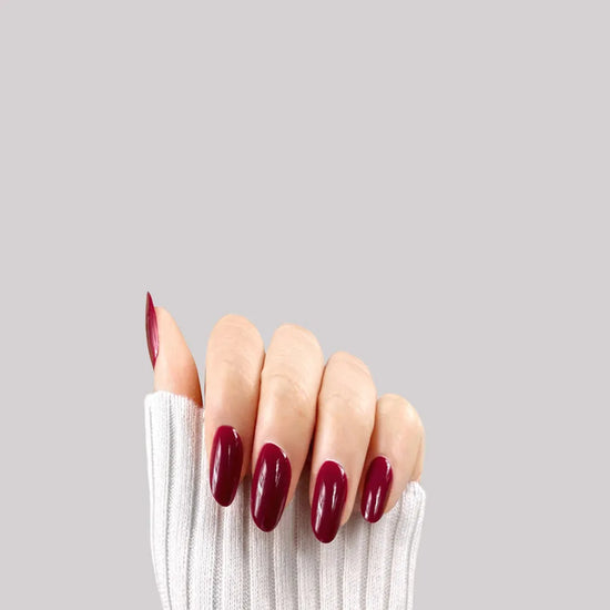 Claw pose showing de blinkie Royal Berry deep cherry red gloss press-on nails in short oval against a grey background with a white ribbed knit sleeve