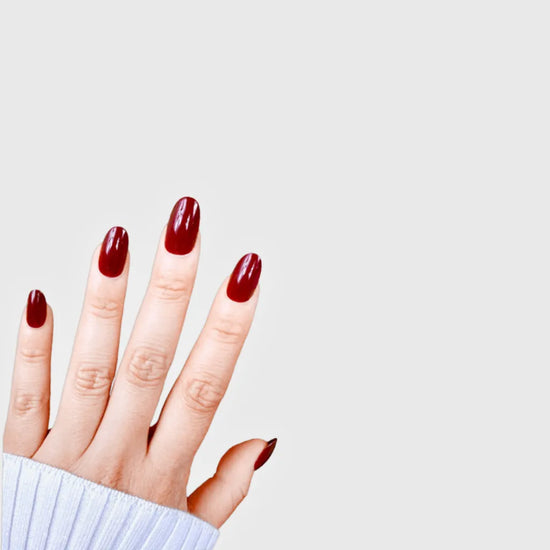 Diagonal hand pose showing de blinkie Royal Berry deep cherry red gloss press-on nails in short oval against a grey background with a white ribbed knit sleeve