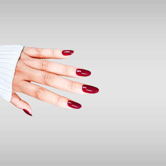 Fingers spread pose showing de blinkie Royal Berry deep cherry red gloss press-on nails in short oval against a grey background with a white ribbed knit sleeve
