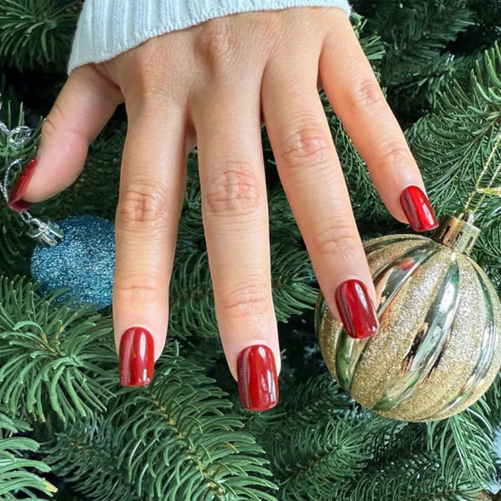 Claw pose showing de blinkie Scarlet Luxe scarlet red mirror chrome press-on nails in short square against pine tree background with gold and blue Christmas ornaments and white knit sleeve