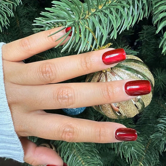 Diagonal hand pose showing de blinkie Scarlet Luxe scarlet red mirror chrome press-on nails in short square holding gold glitter ornament against pine tree background with white knit sleeve