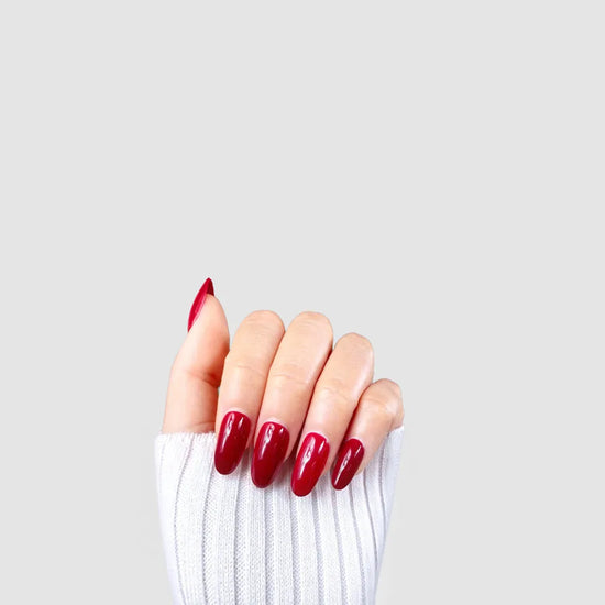 Claw pose showing de blinkie Scarlet Muse classic crimson gloss press-on nails in short oval against a grey background with a white ribbed knit sleeve