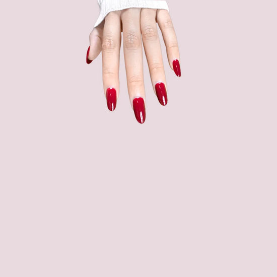 Diagonal hand pose showing de blinkie Scarlet Muse classic crimson gloss press-on nails in short oval against a pink background with a white ribbed knit sleeve