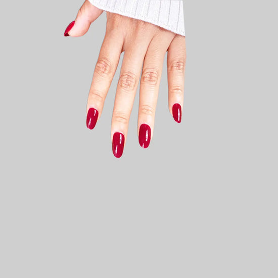 Fingers down pose showing de blinkie Scarlet Muse classic crimson gloss press-on nails in short oval against a grey background with a white ribbed knit sleeve