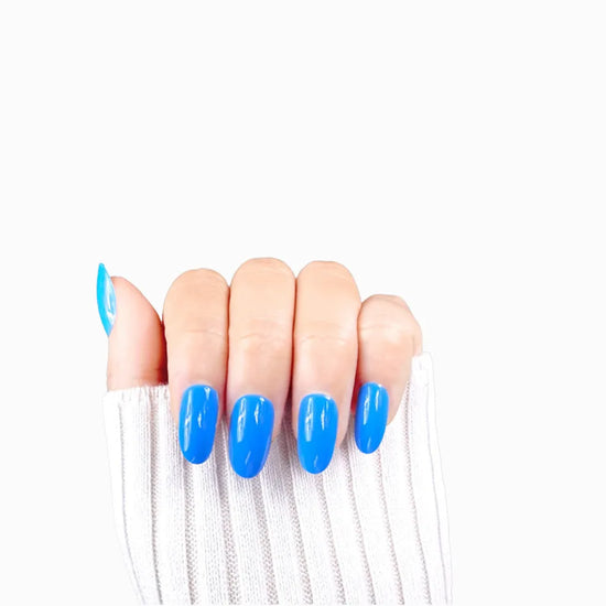Claw pose showing de blinkie Skyline Pop vivid azure blue gloss press-on nails in short oval against a white background with a white ribbed knit sleeve
