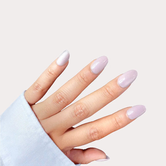 Diagonal spread hand showing Sugar Glaze pale lilac blush silver shimmer frosted short oval press-on nails against off-white background with light blue sleeve