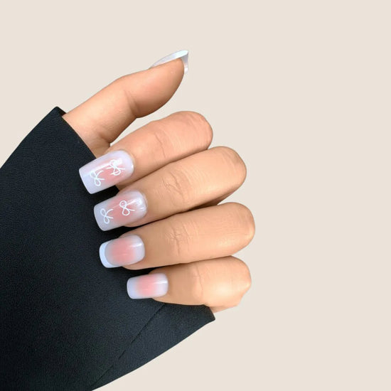 Closed claw pose showing de blinkie Sweetheart Tips rosy blush press-on nails with white bow nail art in short square shape against cream background with black sleeve