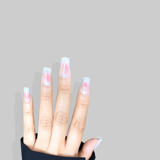 Open downward hand pose showing de blinkie Sweetheart Tips rosy blush press-on nails with white heart and bow nail art in short square shape against light grey background with black sleeve