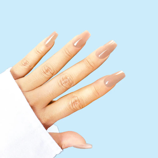 Diagonal spread hand showing de blinkie The Executive Nude warm beige nude press-on nails in short coffin shape against soft blue background with white sleeve