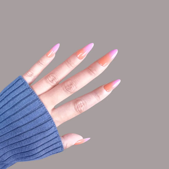 Horizontal spread hand showing de blinkie Tropic Muse coral orange to hot pink ombre press-on nails in medium almond against a soft mauve grey background with a blue ribbed knit sleeve