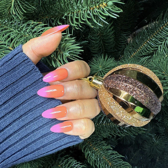 Lifestyle shot showing de blinkie Tropic Muse coral orange to hot pink ombre press-on nails in medium almond holding a gold glitter Christmas ornament against a Christmas tree background with a blue ribbed knit sleeve